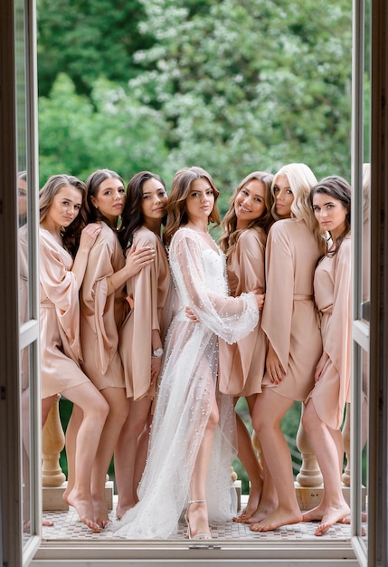 Free photo beautiful bride in elegant wedding dress and bridesmaids dress in beige style robes standing on balcony preparing for wedding ceremony morning of bride