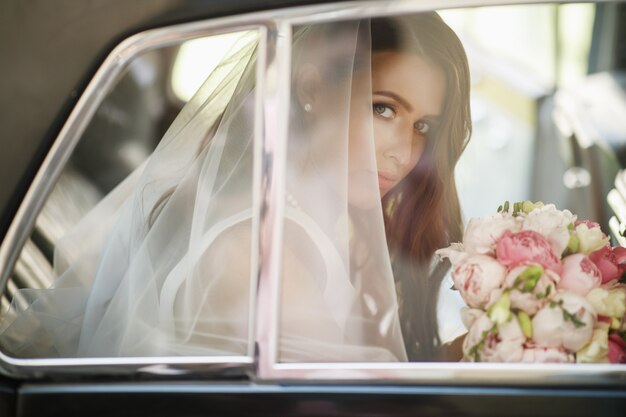 美しい花嫁はレトロ車の結婚式の花束で座って、楽しいです