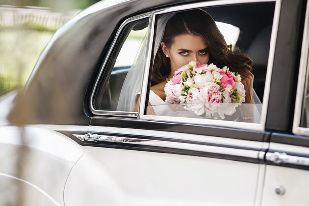 Красивая невеста сидит с свадебным букетом в ретро-машине и весело
