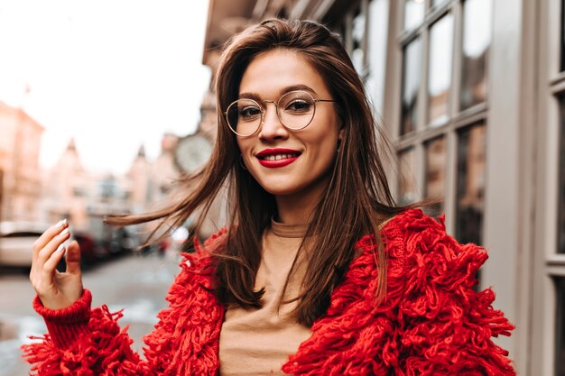 Beautiful brown-eyed woman in glasses coquettishly touches her hair. Woman with red lipstick in bright knitted outfit and headdress walks around city.