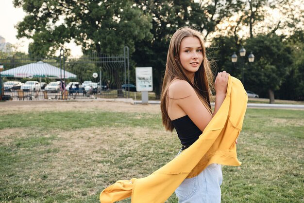 Beautiful brown haired girl in black top and jeans holding yellow shirt in hands while dreamily looking in camera in city park