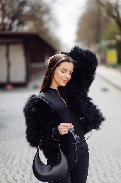 Красивая шатенка стильная девушка в черном внешнем платье. Портрет молодой привлекательной элегантной женщины с длинными волосами на весеннее время на улицах города.