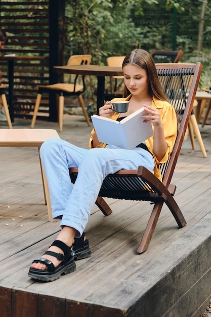 노란색 셔츠와 청바지를 입은 아름다운 갈색 머리 10대 소녀가 도시 공원에 있는 카페의 나무 갑판 의자에 커피 한 잔과 함께 책을 읽고 있습니다.