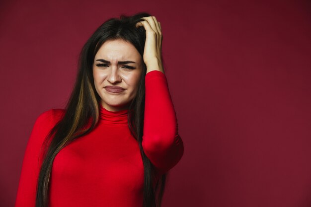 Beautiful brunette baffled caucasian girl dressed in red pullover is scraping her hair