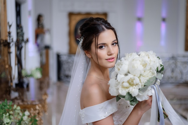 Beautiful brunette caucasian bride is holding bouquet of white peonies and looking straight indoor