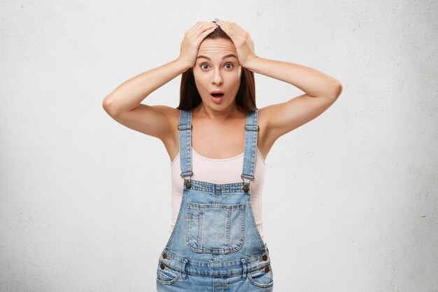 Beautiful brunette female with dark eyes, wearing white T-sirt and denim overalls, keeping hands on head while being shocked to realize something, looking with wide opened mouth and puzzlement