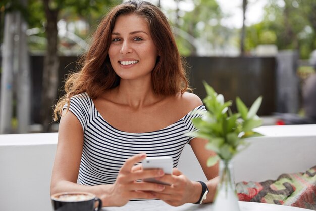 Beautiful brunette female with glad expression and phone in outdoor terrace cafe