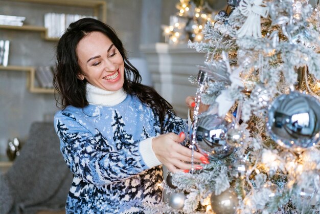 美しいブルネットの女の子は、青い冬のセーターメリークリスマスで人工的なクリスマスツリーを飾ります...