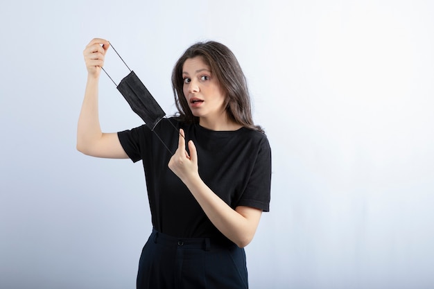 Beautiful brunette girl holding black medical mask over white wall. 