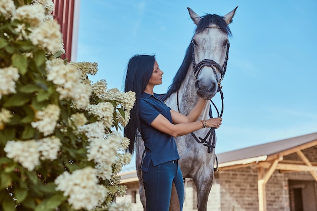 Beautiful brunette girl stroking her gray horse near lilac bushes in the garden.