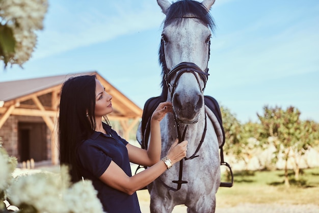 Beautiful brunette girl stroking her gray horse near lilac bushes in the garden.