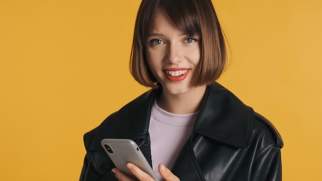 Beautiful brunette girl with bob hair using smartphone happily looking in camera isolated on yellow background