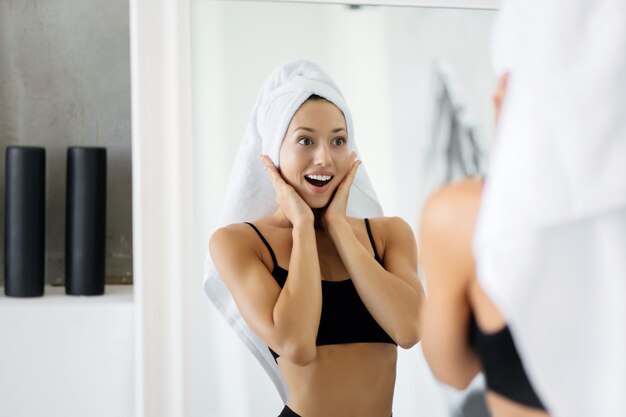 Красивая брюнетка стоит в ванной с полотенцем на голове перед зеркалом. Утренние процедуры и уход за здоровьем