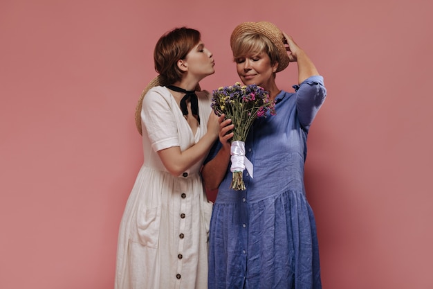 Beautiful brunette lady in white clothes blowing kiss and posing with old woman in blue dress and hat with wildflowers on isolated backdrop.