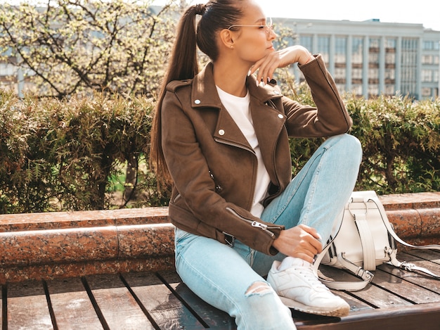 Bellissima modella bruna vestita in abiti hipster estate giacca e jeans ragazza alla moda, seduto sulla panchina in strada