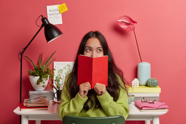 Beautiful brunette pensive mixed race female student learns information from textbook, covers half of face with red diary