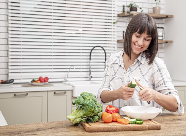 Beautiful brunette smiling and preparing salad on blurred background of kitchen interior.