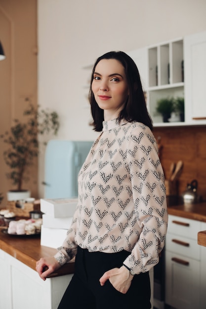 Beautiful brunette standing in the kitchen. patterned blouse and dark trousers or jeans standing in the modern kitchen and looking away thoughtfully..