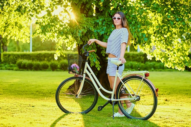 Beautiful brunette stands on the green lawn with bicycle in park