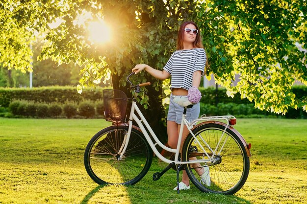 Beautiful brunette stands on the green lawn with bicycle in park