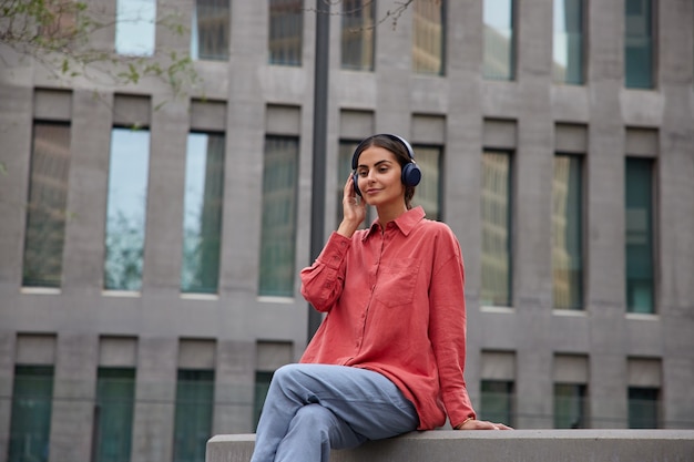 Beautiful brunette woamn sits outdoors listens music or audio podcast via wireless headphones enjoys leisure pastime dressed in red shirt and trousers takes break after strolling