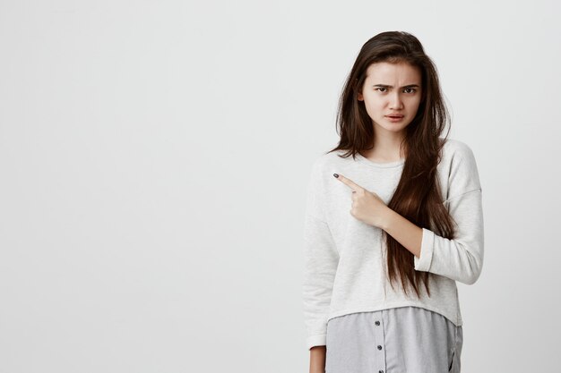 beautiful brunette woman in casual clothes pointing with index finger at gray blank wall having dissatisfied look, frowning, with negative expression of face