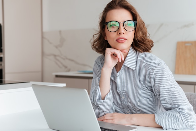 beautiful brunette woman in glasses holding her chin and looking aside while sitting at workplace