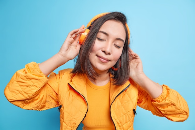  beautiful brunette woman has eyes closed wears wireless headphones listens music tilts head dressed in orange jacket isolatedover blue wall. People lifestyle hobby concept