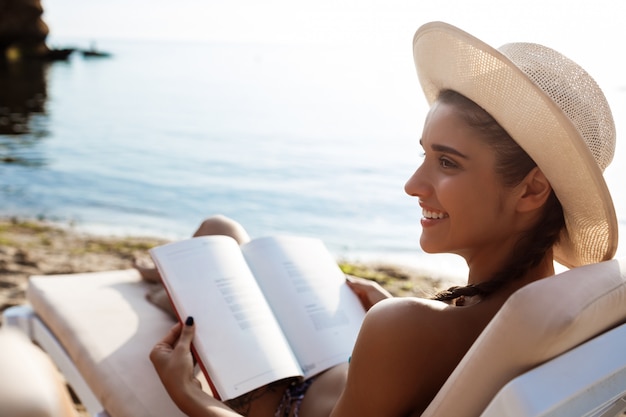 Free photo beautiful brunette woman in hat reading book, lying at beach