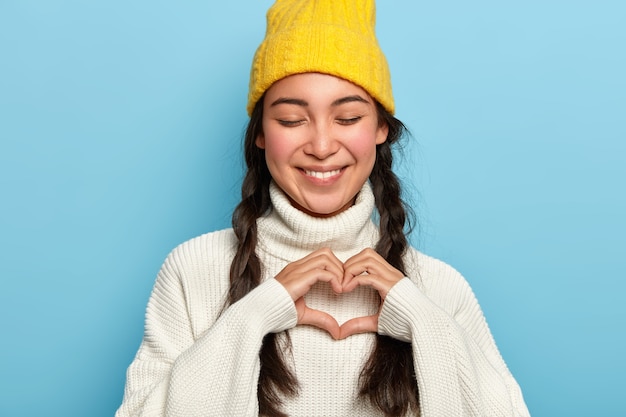 Beautiful brunette woman keeps eyes closed with pleasure, makes heart gesture, expresses her true love, dressed in casual white sweater