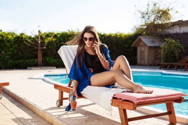 Beautiful brunette woman speaking on phone, sitting near swimming pool