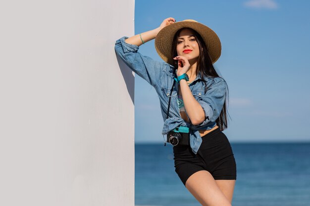 Beautiful brunette woman with figure in shorts and straw hat posing on the beach.