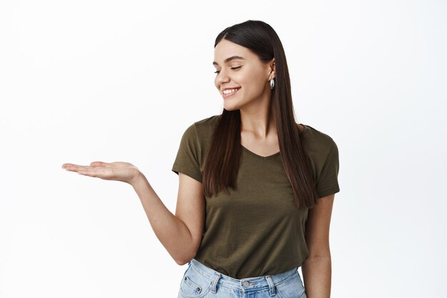 Beautiful brunette woman with natural make up wearing green tshirt holding your logo product on palm looking at empty hand on copy space white background