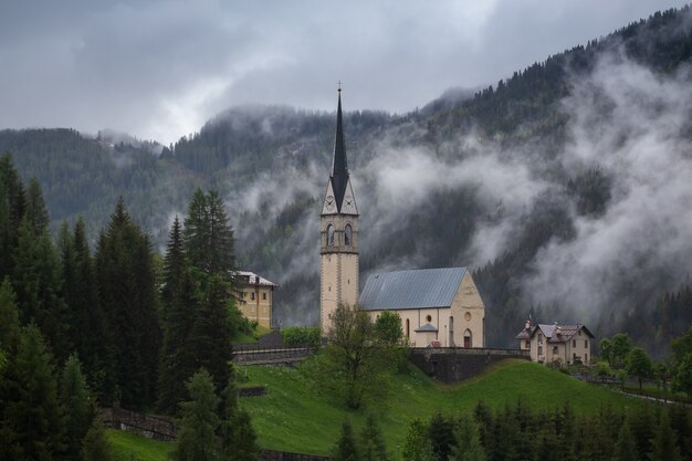 Beautiful buildings in a foggy green forest with dense trees and mountains in Dolomites, Italy