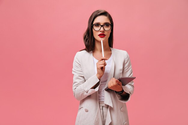 Beautiful business lady in eyeglasses and modern suit thoughtfully posing with computer tablet on isolated pink background.