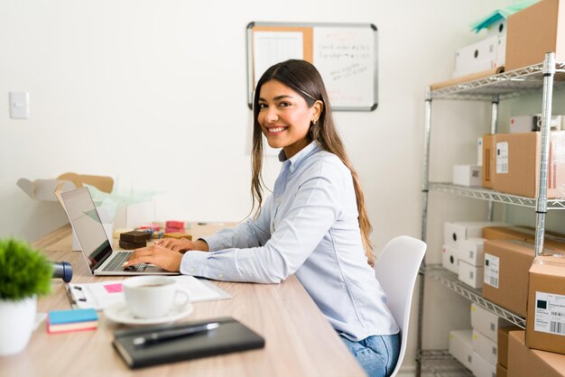 Красивая владелица бизнеса улыбается и смотрит в глаза, работая за своим офисным столом в своей онлайн-стартапе
