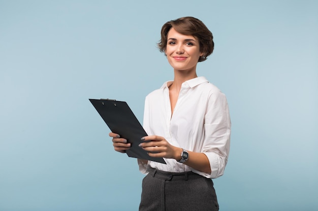 Красивая деловая женщина с темными короткими волосами в белой рубашке держит в руках черную папку, радостно глядя в камеру на синем фоне