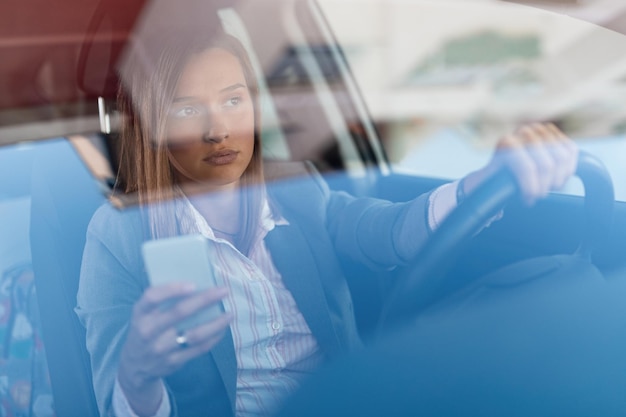 운전을 하고 운전석에서 휴대전화를 사용하는 아름다운 여성 사업가가 유리를 통해 전망을 볼 수 있습니다