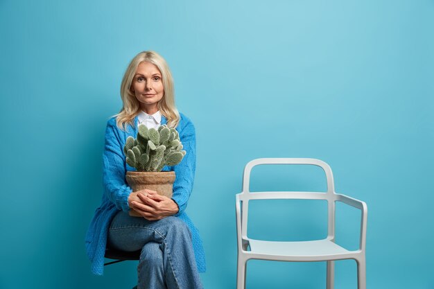 Beautiful calm confident wrinkled European woman feels nostalgia melancholy holds with potted cactus poses with empty chair