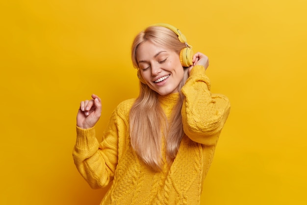 Beautiful carefree blonde woman dances keeps arm raised relaxes with pleasant music from headphones