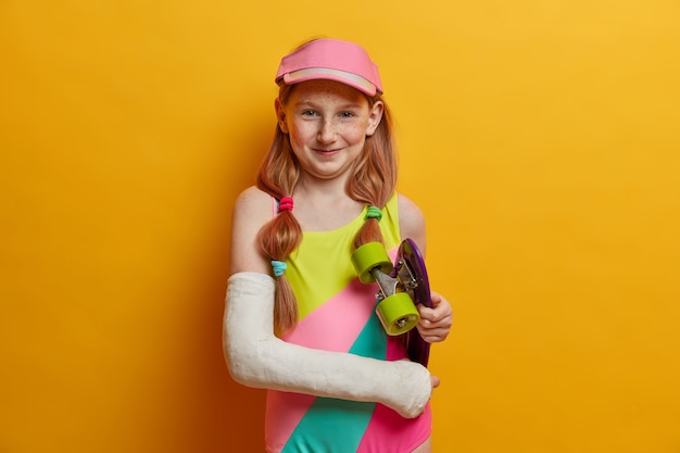 Beautiful carefree girl with two pony tails, glad after riding skateboard, wears cast on broken arm, dressed in bathing suit and cap, spends free time in skatepark, isolated on yellow wall