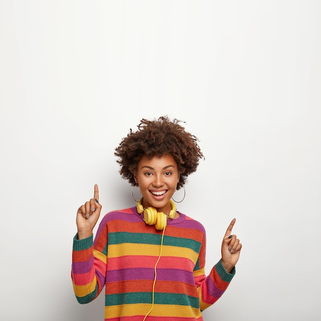 Free photo beautiful carefree hipster girl with afro hairstyle, moves against white wall, points upwards, says your text here, uses yellow headphones for listening favourite music, wears striped colored jumper