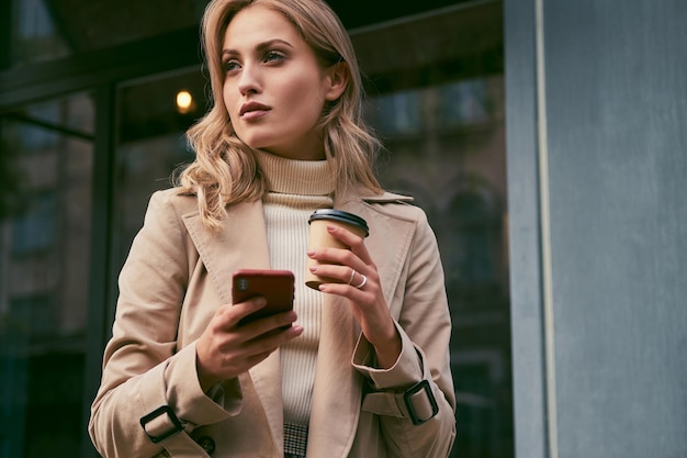 Красивая повседневная блондинка в плаще с кофе на вынос и мобильным телефоном задумчиво смотрит в сторону на улице