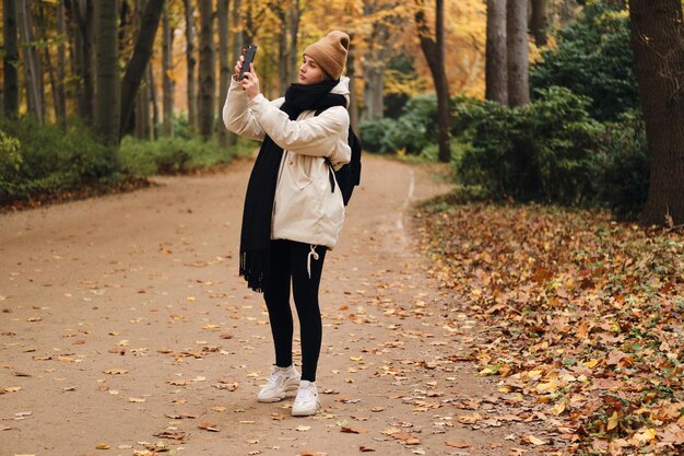 秋の公園を歩いて携帯電話で写真を撮る美しいカジュアルな女の子