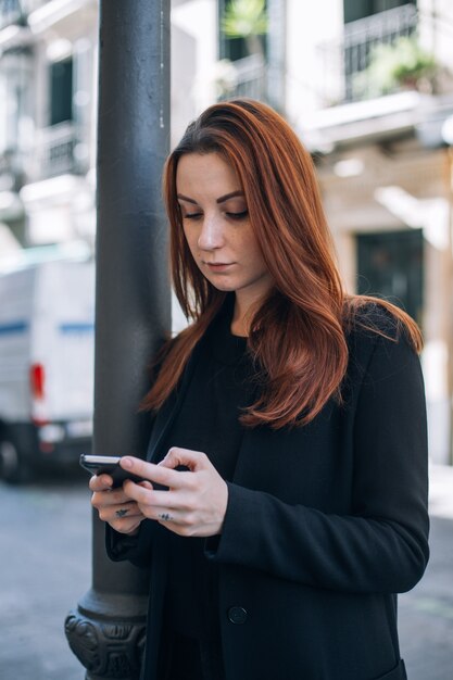 赤い髪とナチュラルメイクの美しいカジュアルな女性が通りに立って、スマートフォンでテキストやチャットをします