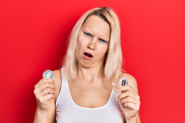 Free photo beautiful caucasian blonde woman holding virtual currency ethereum coin and bitcoin in shock face looking skeptical and sarcastic surprised with open mouth