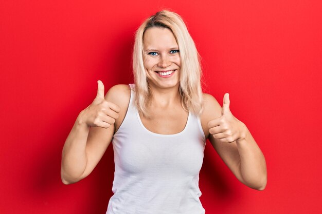 Beautiful caucasian blonde woman wearing casual white t shirt success sign doing positive gesture with hand, thumbs up smiling and happy. cheerful expression and winner gesture.