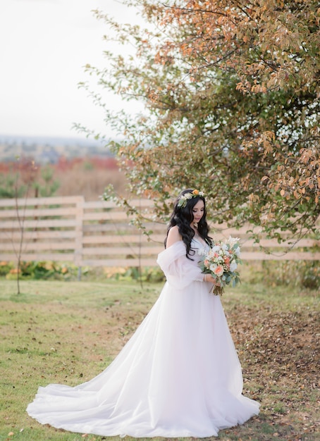 暖かい秋の日に木の近くの乾いた草の上にウェディングブーケを持つ美しい白人花嫁が立っています。