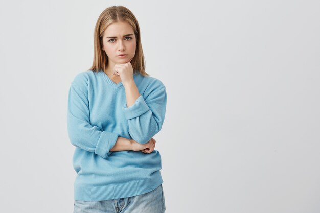 Beautiful caucasian dissatisfied female with straight fair hair wearing casual clothes looking  with anger, holding her hands under her chin, dissatisfied with actions of her boyfriend.