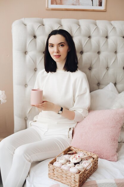 Beautiful caucasian female with short dark hair in a white cozy sweater holds a yoummy marshmallow and smiles